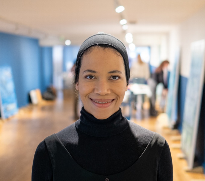 Eve stands in a gallary space with blue walls and light brown floor. Paintings lean up against the walls behind her. She faces the camera, wearing her hair pushed back in a wide headband, and a black, roll-necked top.