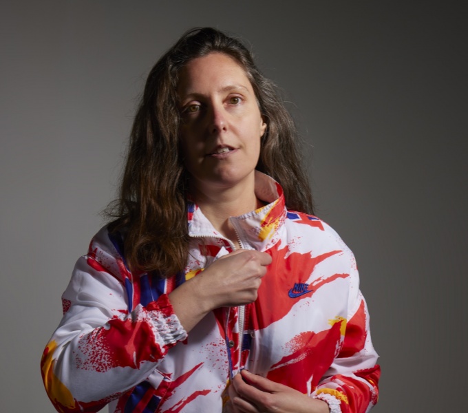 A portrait photograph of Louise Ashcroft. A whaite womal with long brown hair wearing a white jacket with abstract paint effect design in bright red, blues and yellow. 