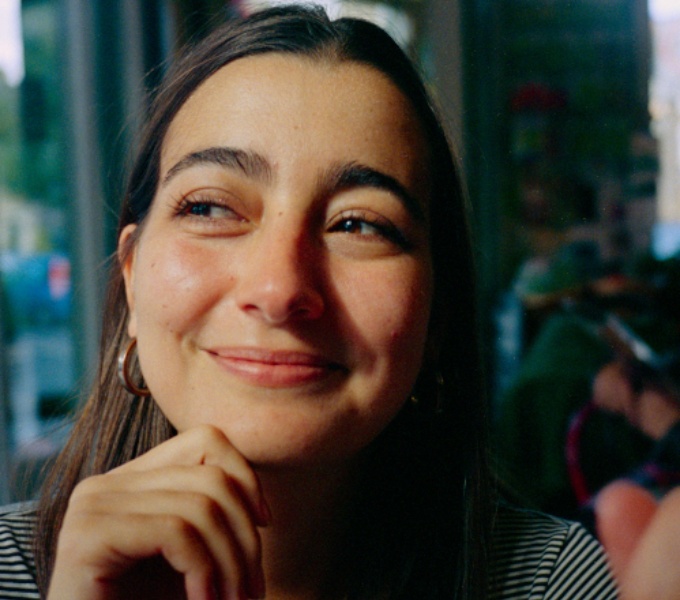 A head and shoulders, colour photographic portrait of Mariana Lemos. Mariana has shoulder length brown hair, dark brown eyebrows and brown eyes. She has tanned skin. She wears gold hooped earrings, a gold cross pendant on a chain around her neck.