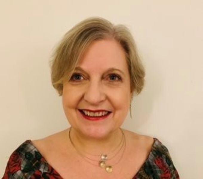 A portrait photograph of Morag. She wears a red and black dress and a gold necklace with circular disc design. She has short bobbed, blonde coloured hair and red lipstick. He gaze is to the left of the camera and she is smiling.  