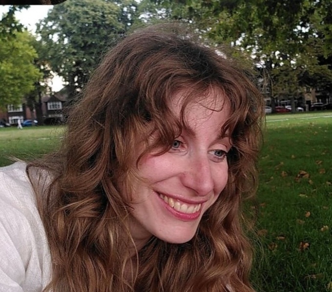 Headshot of Nathalie, a white woman with blue eyes and long dark blonde hair. She wears a white top and is seated on the grass of a park. Behind her in the distance are houses. She is smiling broadly.