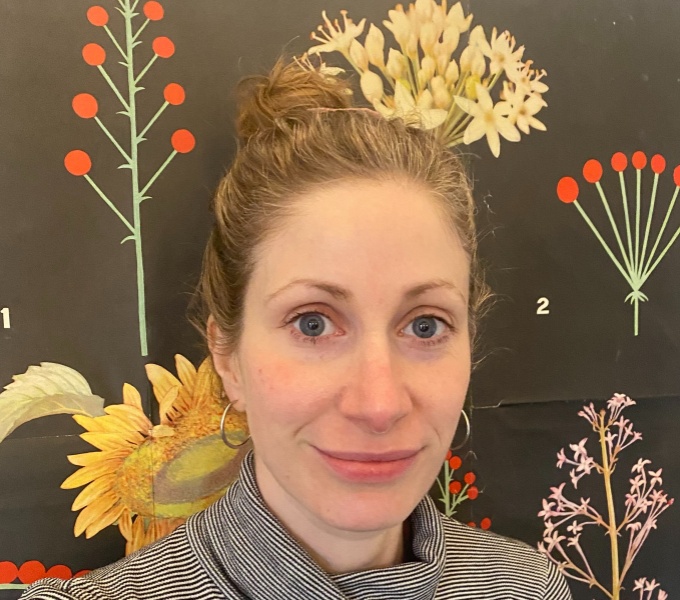 Headshot of Nathalie, a white woman with blue eyes and dark blonde hair tied up into a bun. She is looking directly at the camera and is smiling. She is wearing a striped polo neck and silver hoop earrings.