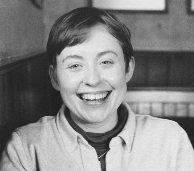 Rachel is a white woman in her late twenties with short hair. In this black-and-white photograph, she is wearing a high-necked top under a thick, buttoned overshirt. She is laughing at the camera and is pictured seated in the interior of her favourite pub