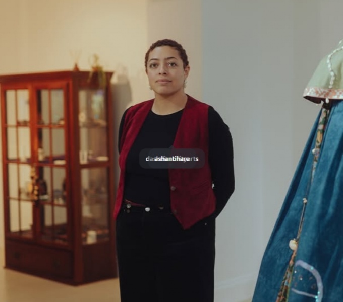Curator Ashanti Hare stands in a white-walled gallery space. They are captured from the knees upwards. In the background to the left is a wooden glazed display cabinet, and in the foreground to the right is a blue, green and cream textile costume.