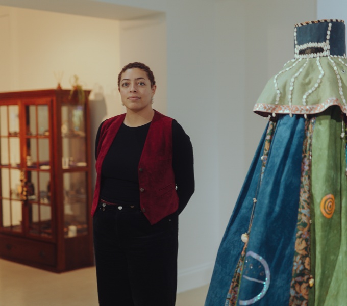 Curator Ashanti Hare stands in a white-walled gallery space. They are captured from the knees upwards. In the background to the left is a wooden glazed display cabinet, and in the foreground to the right is a blue, green and cream textile costume.