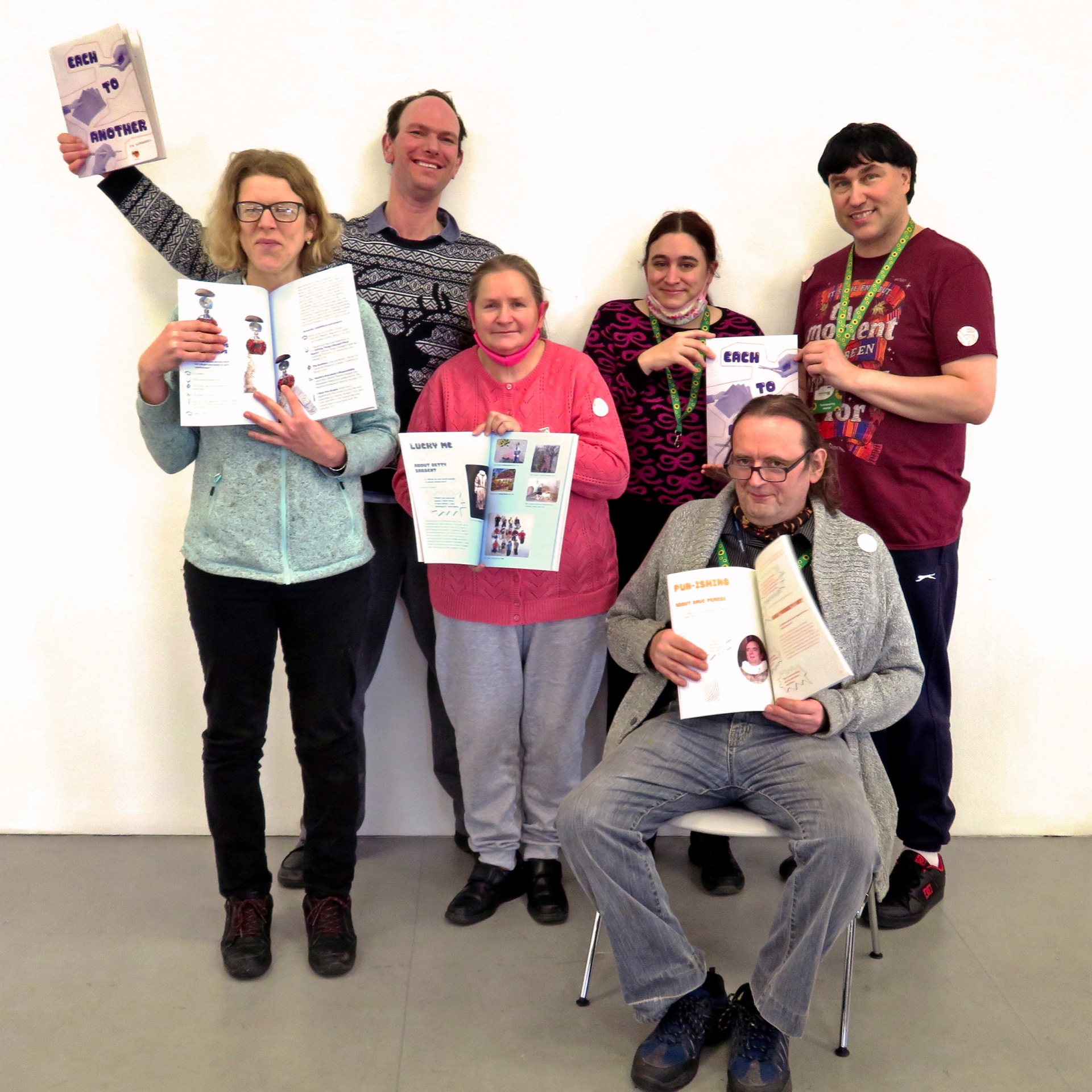 Six AIM artists stand together and hold up copies of their new publication. There are three women and three men. The man in the foreground is seated.