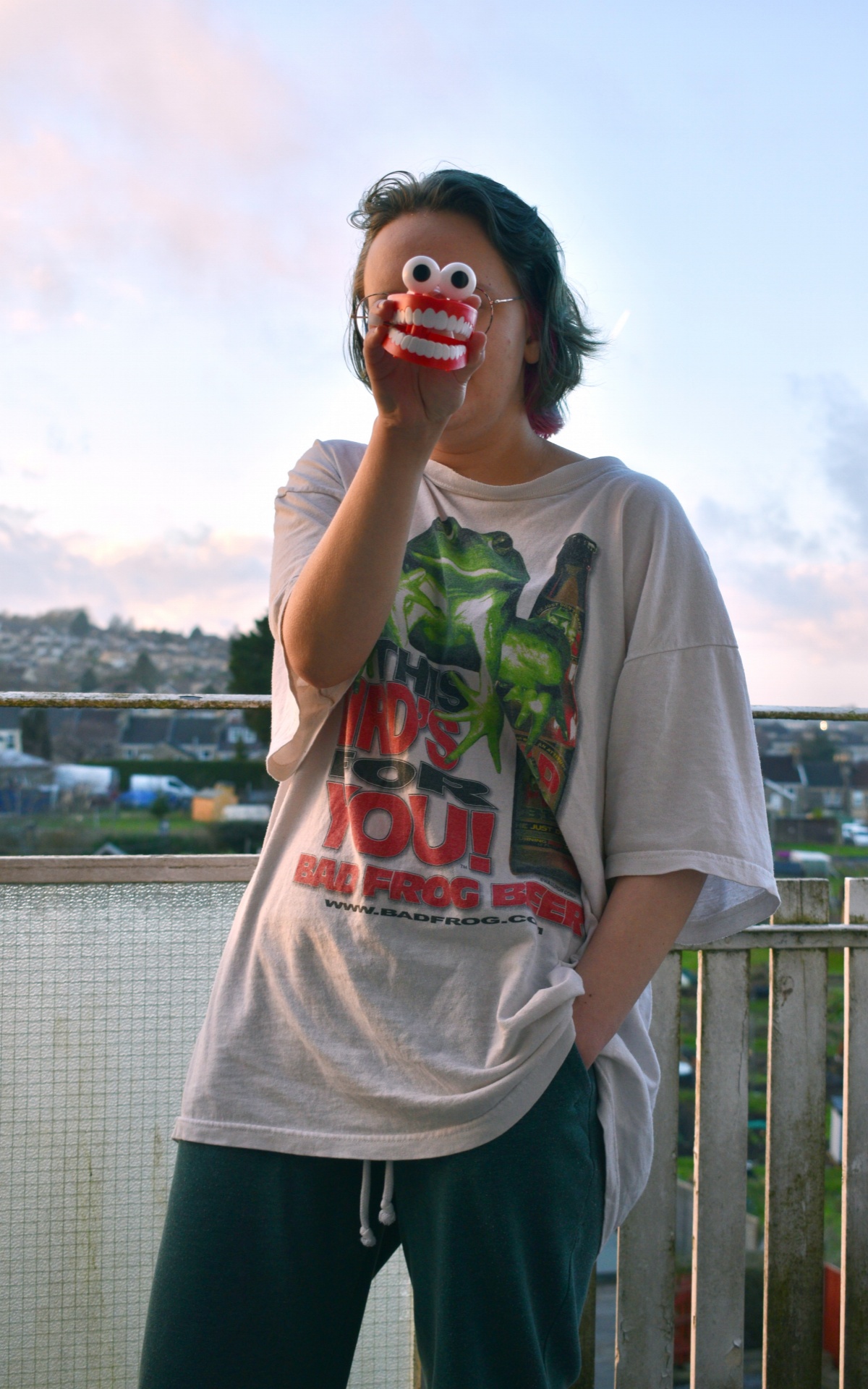 Alice stands next to the waters edge in an oversized t-shirt and jeans. One hand is in her pocket  whilst the holds a large set of toy wind up teeth in front of her face, obscuring her features.