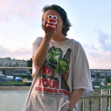 Alice stands next to the waters edge in an oversized t-shirt and jeans. One hand is in her pocket  whilst the holds a large set of toy wind up teeth in front of her face, obscuring her features.