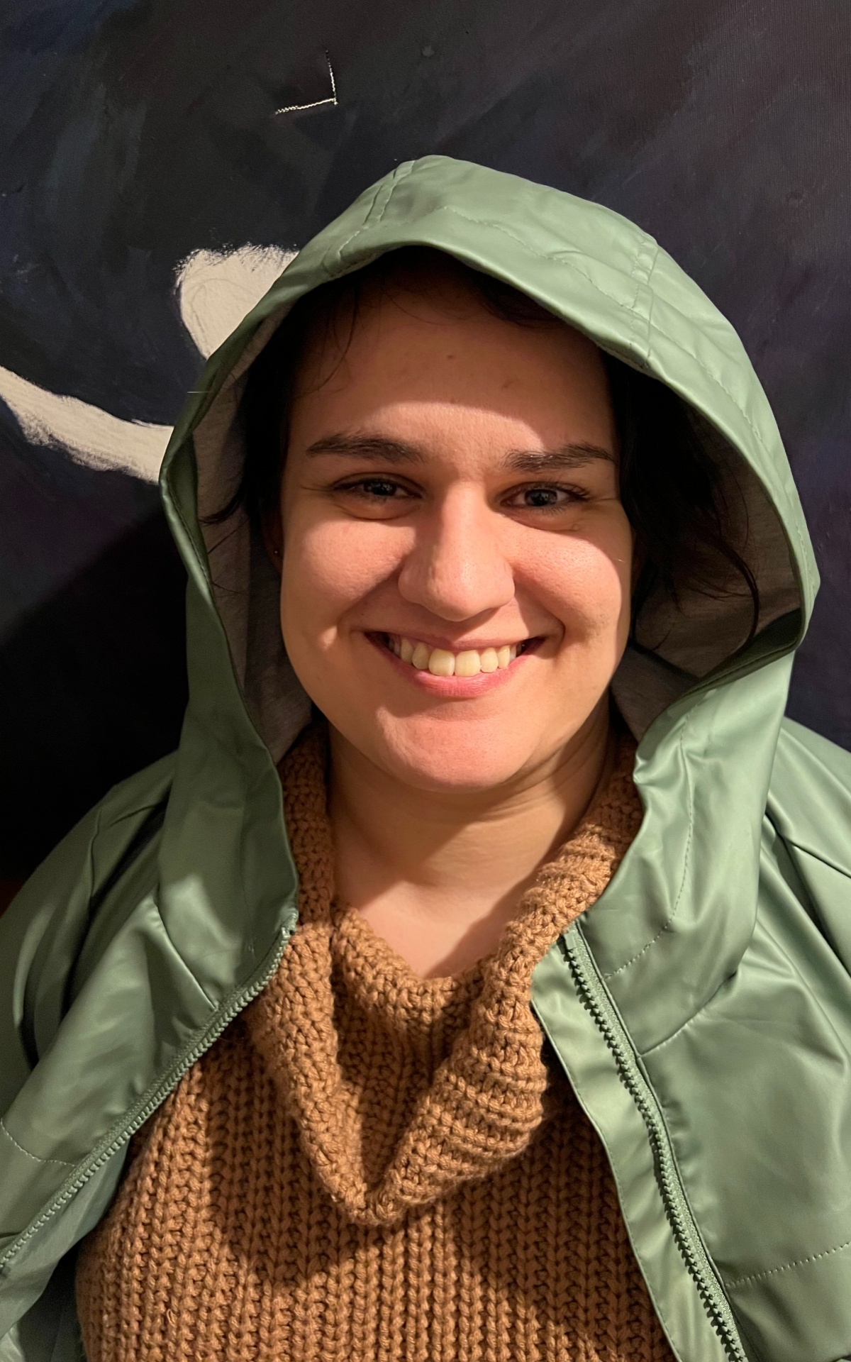 Anahita smiles at the camera. She wears a sage green rain mac with the hood pulled loosely over her head, with a sand coloured jumper underneath.