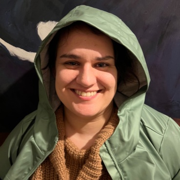 Anahita smiles at the camera. She wears a sage green rain mac with the hood pulled loosely over her head, with a sand coloured jumper underneath.