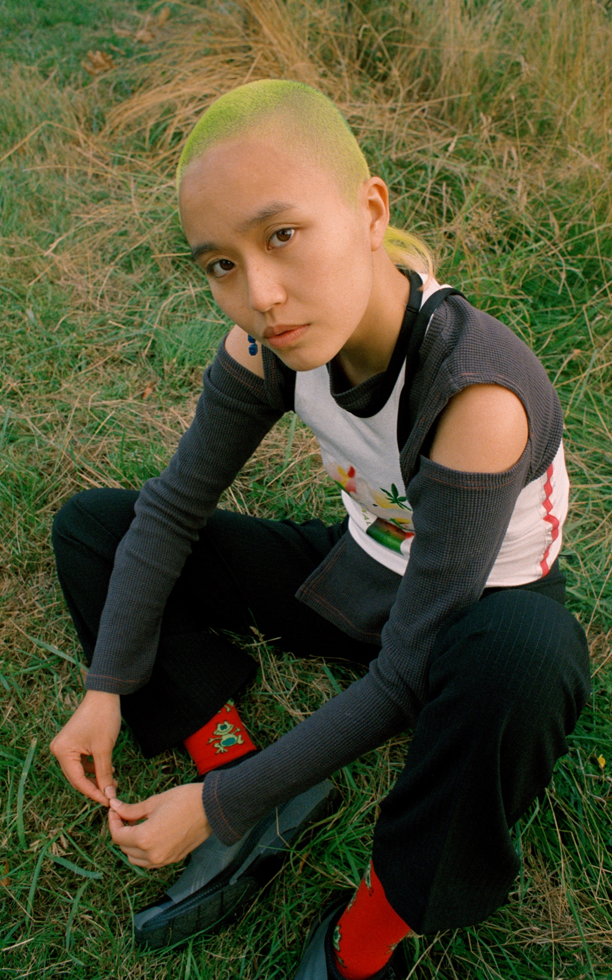 April sits on the grass crosss legged, ad looks upwards towards the camera. She has a shaved hair cut which is dyed a lime green colour and wears black trousers, a long sleeved grey and white top.