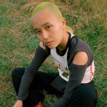 April sits on the grass crosss legged, ad looks upwards towards the camera. She has a shaved hair cut which is dyed a lime green colour and wears black trousers, a long sleeved grey and white top.