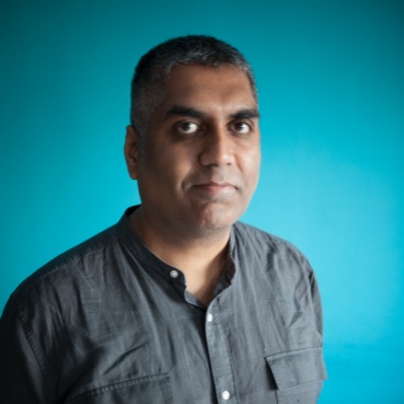 Ashok stands facing the camera against a turquoise background. He has short hair and wears a grey shirt. . He looks directly at the camera.