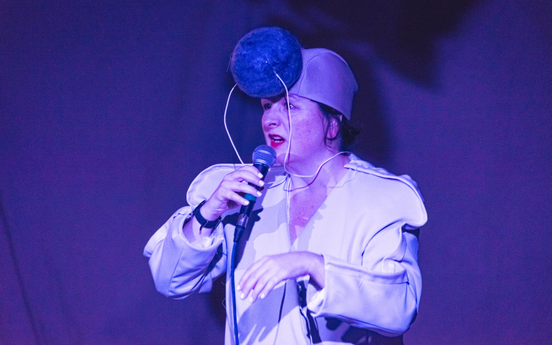 Nicola wears a light coloured jacket and a cap with sound equipment attached to the front of her head. It is shaped like a large pom pom. She holds the microphone to her lips as she addresses and audience. The room is lit with a purple, blue lighting. 