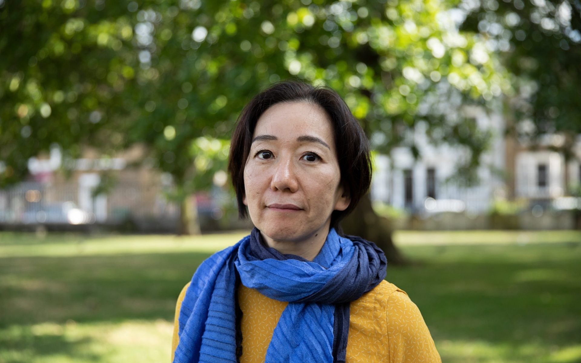 artist Chisato Minamimura standing in a green space outdoors. She wears a mustard jumper and a two tone light and dark blue scarf.