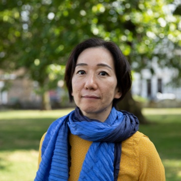 artist Chisato Minamimura standing in a green space outdoors. She wears a mustard jumper and a two tone light and dark blue scarf.