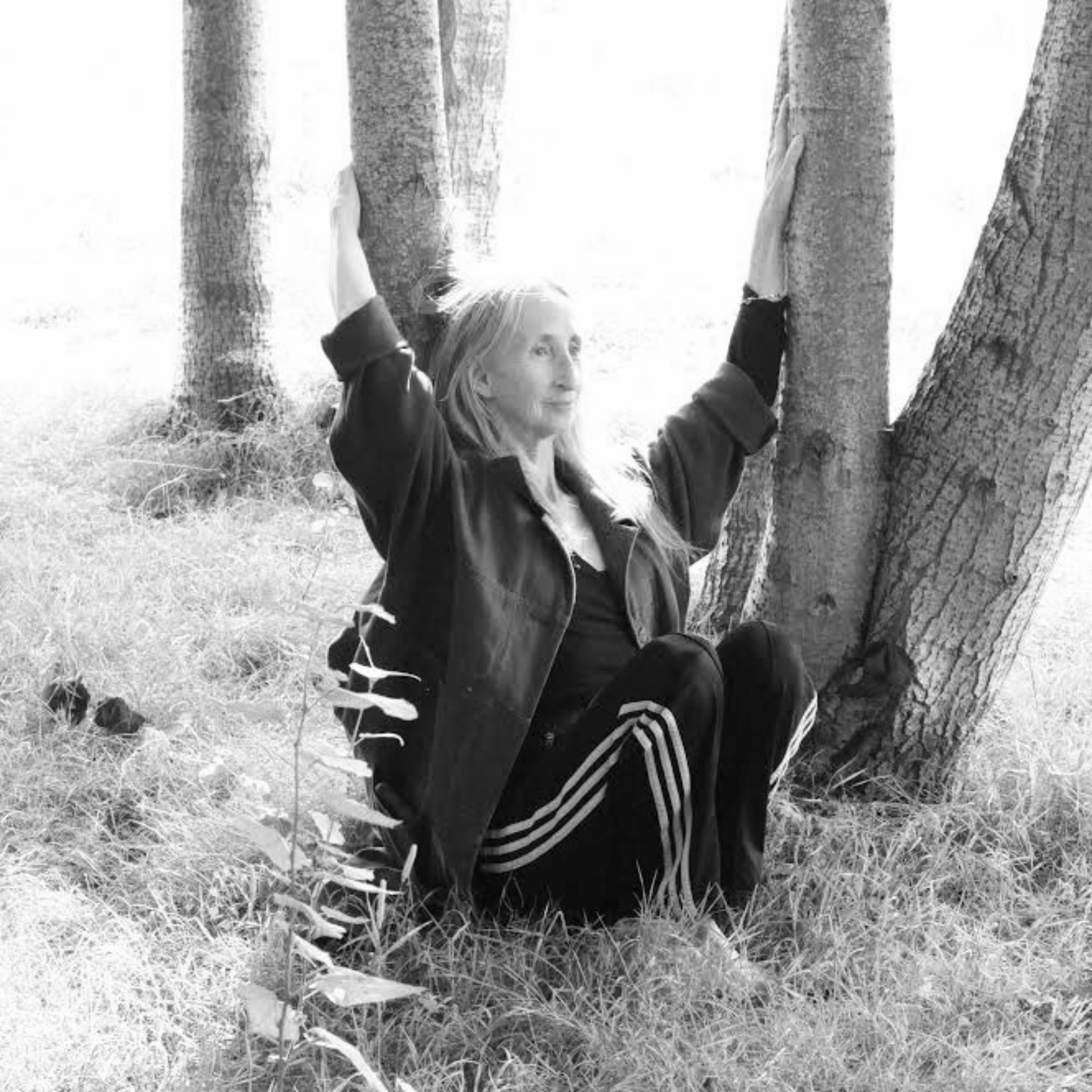 A black and white photograph of Christina sat on grass amongst young trees. She wears black tracksuit bottoms with 3 white stripes running up the side with a dark coloured jacket. She has her arms upstretched and holds the trunks of two trees.