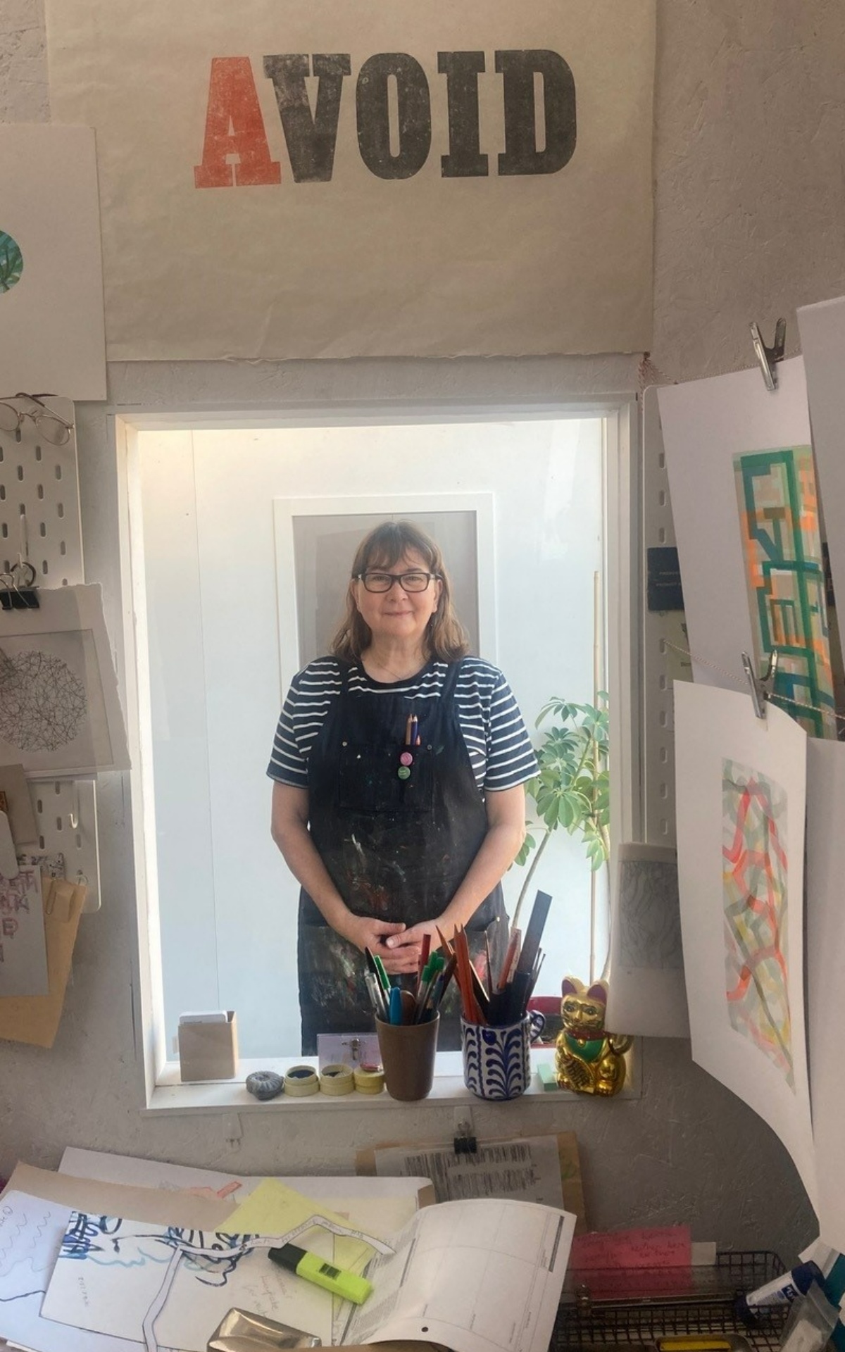 Emma stands behind an internal windowlike, hatch. She wears a blue and white short sleeved t-shirt, a navy blue apron and dark rimmed glasses. Her hands are clasped. In front of the hatch, on walls and table, are pieces of artwork, paper and equipment. 