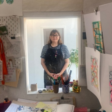 Emma stands behind an internal windowlike, hatch. She wears a blue and white short sleeved t-shirt, a navy blue apron and dark rimmed glasses. Her hands are clasped. In front of the hatch, on walls and table, are pieces of artwork, paper and equipment. 