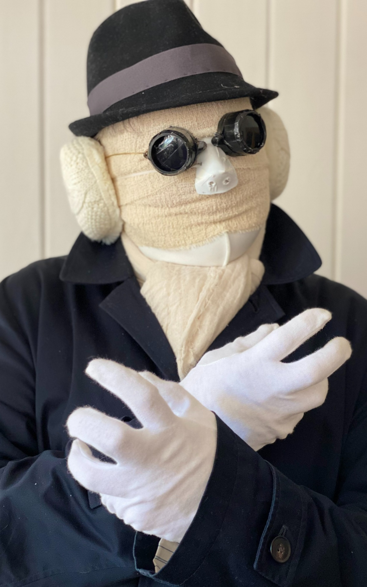 A person with a fully covered head in bandages including ears. They wear a black ‘pork pie’ style hat, blacked out goggles and a black mac style coat. Their white gloved hands are crossed in front of them below their neck. 