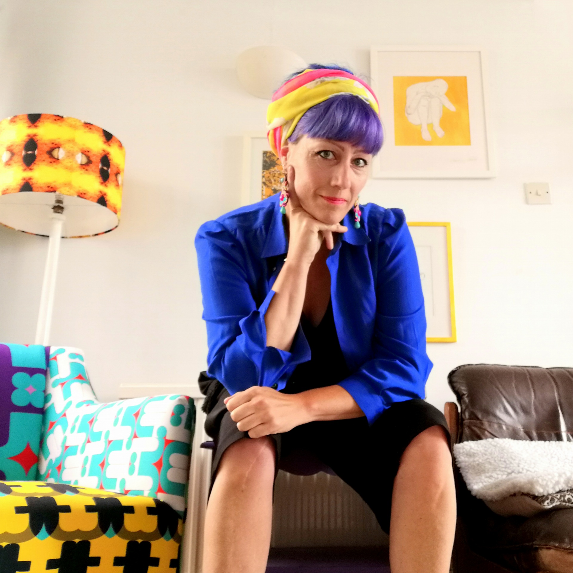 Kristina sits on a stool in a bright white room with retro accessories in orange and yellow. She wears a blue shirt, short black trousers and a scarf around her head. She rests her chin on her right hand.
