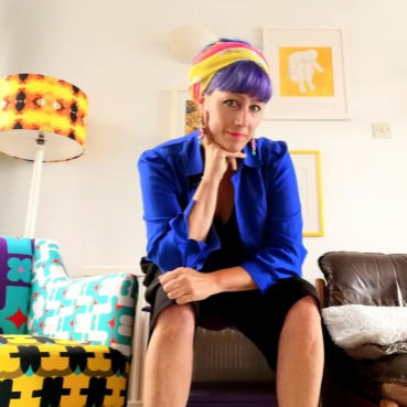 Kristina sits on a stool in a bright white room with retro accessories in orange and yellow. She wears a blue shirt, short black trousers and a scarf around her head. She rests her chin on her right hand.