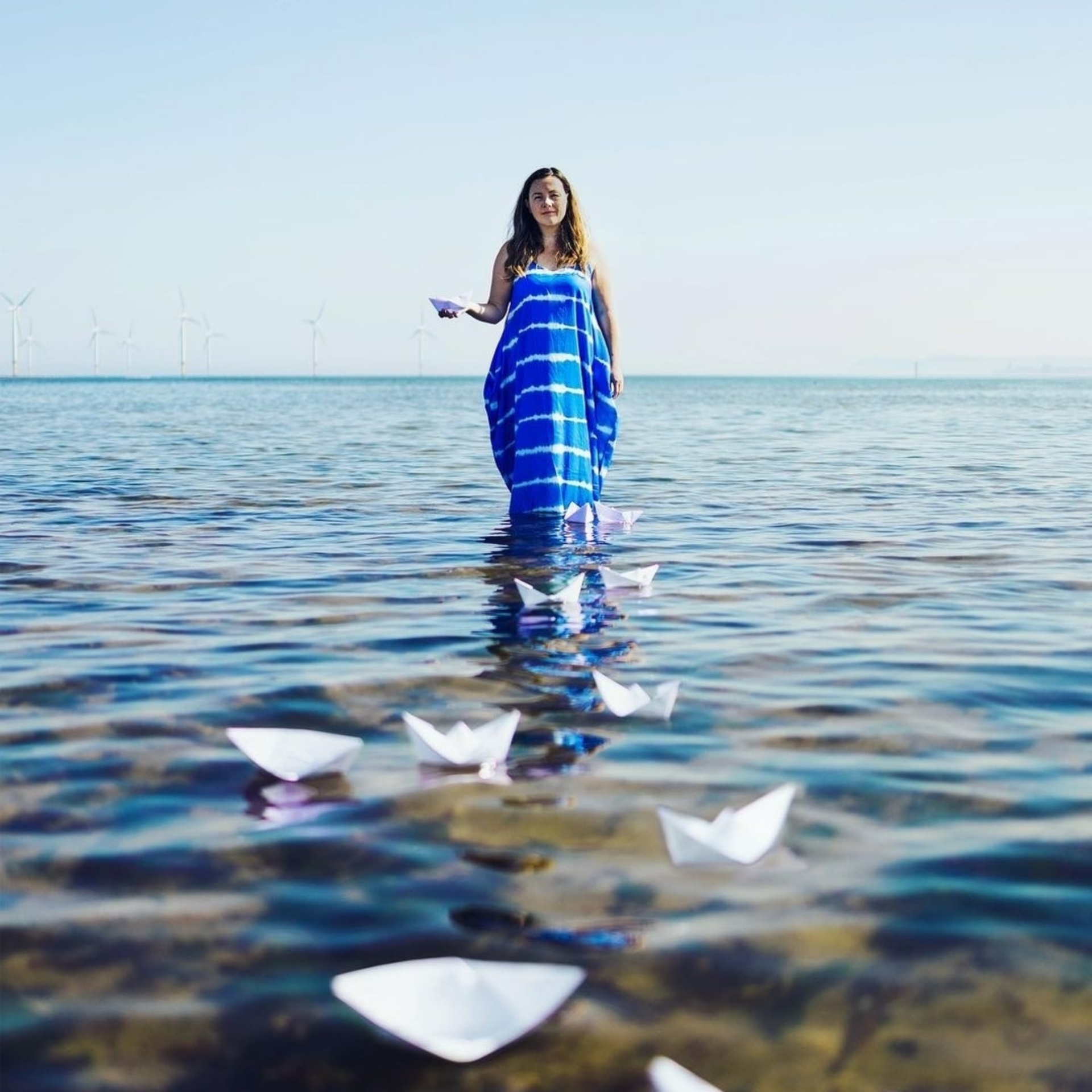 Lisette stands on a rocks surrounded by sea water. She wears her hair down and a long blue dress. Her right hand is held out in front of her, floating on the water a several white origami paper boats.