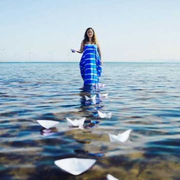 Lisette stands on a rocks surrounded by sea water. She wears her hair down and a long blue dress. Her right hand is held out in front of her, floating on the water a several white origami paper boats.