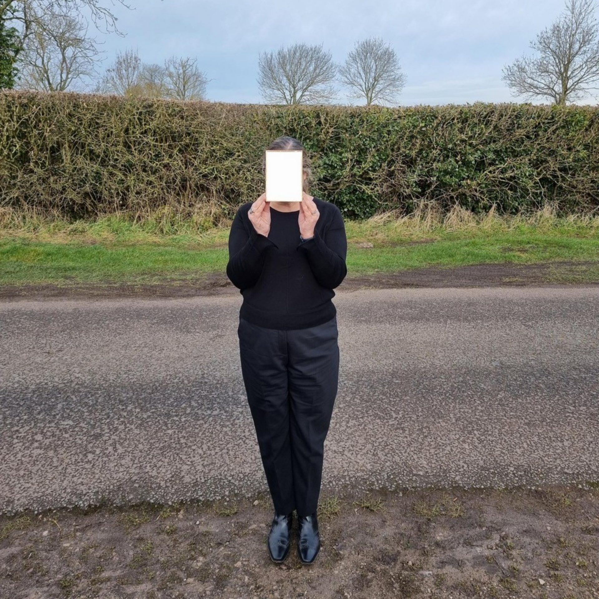 Mianam stands on a roadside, a green hedge behind. She wears black shoes, trousers and jumper and holds a piece of cream coloured card up to her face, hiding her from view.  