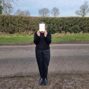 Mianam stands on a roadside, a green hedge behind. She wears black shoes, trousers and jumper and holds a piece of cream coloured card up to her face, hiding her from view.  