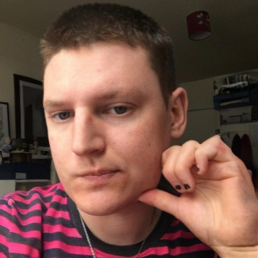 A close up portrait photo of Luca. They have short cropped hair and wears a red and black striped t-shirt and black painted nails and silver chain. 