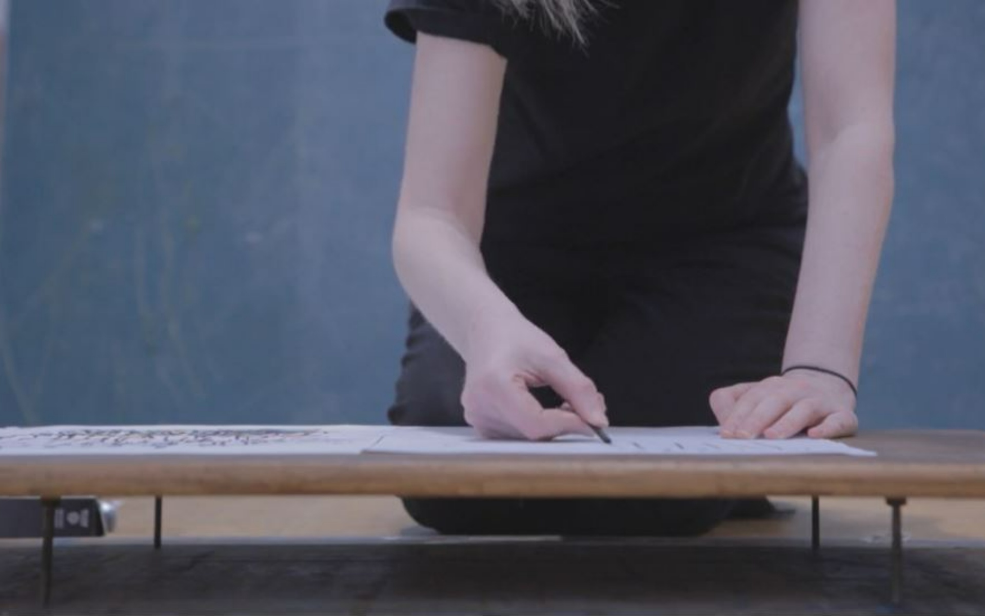 A cropped image showing the arms and hands of a person working on a raised wooden board at a table as they draw. They wear a black t-shirt with a black hair band around their left wrist and hold a piece of charcoal in their right hand. 