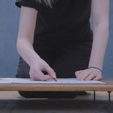 A cropped image showing the arms and hands of a person working on a raised wooden board at a table as they draw. They wear a black t-shirt with a black hair band around their left wrist and hold a piece of charcoal in their right hand. 