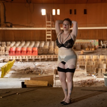 Stav stands on the stage of a run down, dilapidated theatre, arms up and hand resting on their head. They wear black underwear and stockings with a pale pink suspender belt.