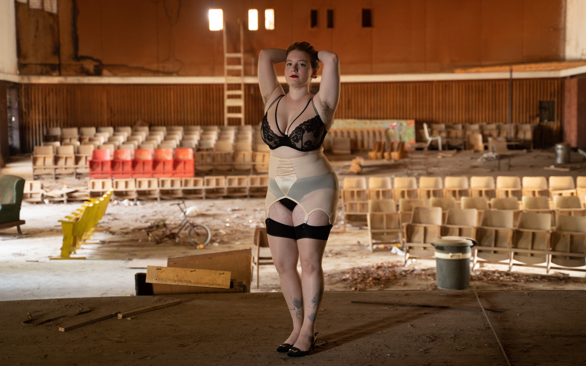 Stav stands on the stage of a run down, dilapidated theatre, arms up and hand resting on their head. They wear black underwear and stockings with a pale pink suspender belt.