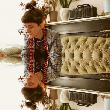 Stephanie wears a grey jacket, dangly earrings and hair tied back in a high bun.  Steph and her surroundings are reflected as if in a mirror in the lower half of the image.