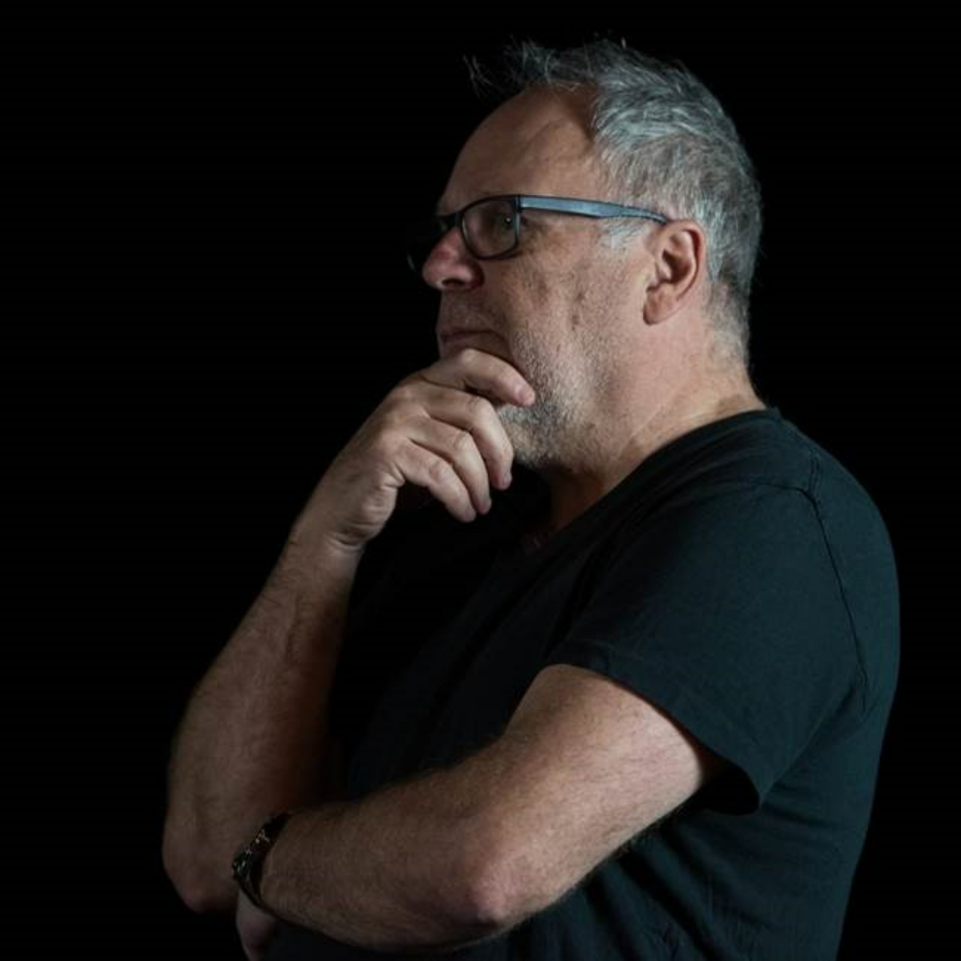 A side image of Terry Smith. He has short grey hair and wears a black t-shirt. He faces to the left, holding his right hand up to his chin in thought, his left arm across his body.
