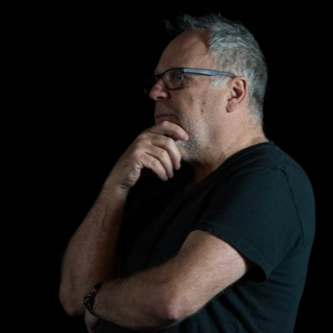 A side image of Terry Smith. He has short grey hair and wears a black t-shirt. He faces to the left, holding his right hand up to his chin in thought, his left arm across his body.
