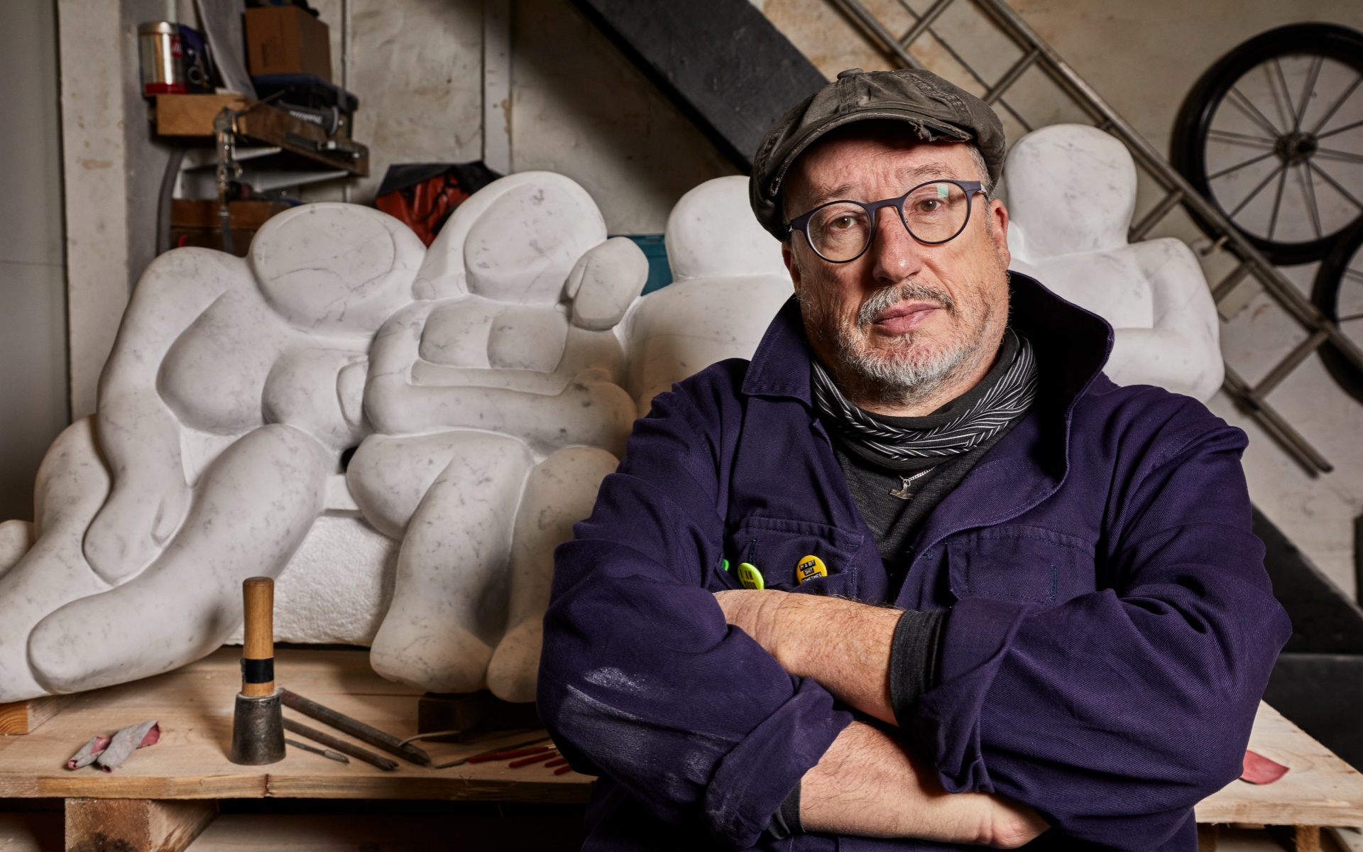 Tony Heaton in front of a large pale abstract sculpture. He wears a flat cap and glasses and dark blue overalls. His arms are folded as he looks at the camera.