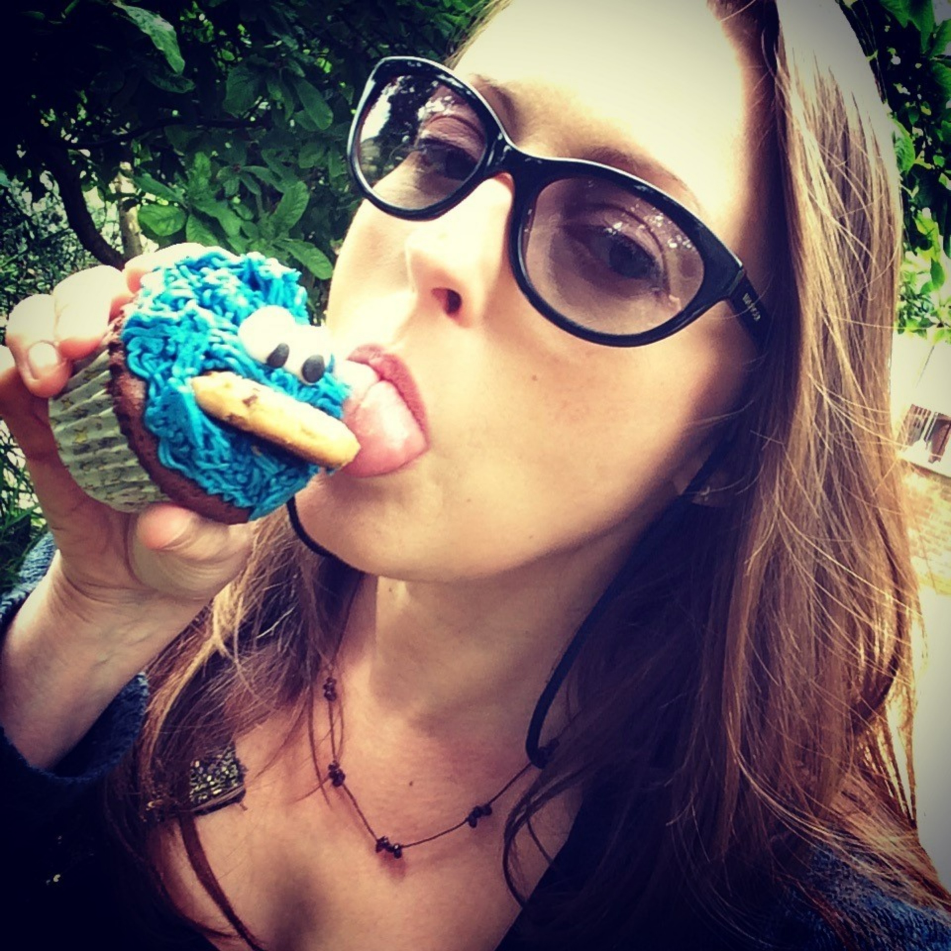 A close up portrait image of Caroline's face. She has long brown hair and wears sun glasses. Her head is tilted to the side as she looks at the camera and takes a bite from a cupcake with turquoise blue icing. 