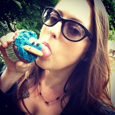A close up portrait image of Caroline's face. She has long brown hair and wears sun glasses. Her head is tilted to the side as she looks at the camera and takes a bite from a cupcake with turquoise blue icing. 