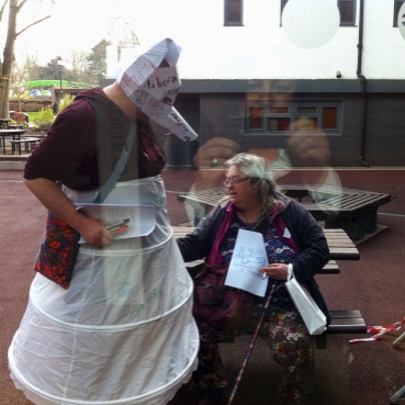 gobscure standing outdoors wearing a structured white petticoat which has been written on. The artist wears a 3D paper head mask with a large pointed nose that obscures their face. A seated woman appears to adjust the fabric.