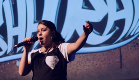 A young woman stands with her left arm outstretched as she speaks into a microphone. She wears a clack and white t-shirt with her dark hair tied back. She stands in front of a concrete wall that is covered with graffiti in blue hues. 