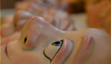 A mask lying flat on a table, seen in profile, creamy white with staring eye looking upwards, nose seen in relief and red lips. in the background there are other masks blurred, lying in lines