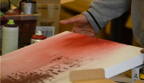 A white canvas with red spray paint at one end, with diffused red colour flowing down over the canvas, meeting a dark brown imprint, is held up slightly at one end by hands of an unseen person. In the background are other cans of spray. 