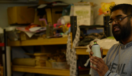 Mohammed stands at the right side of the photo in a white t-shirt with a spray can in his right hand, in the back ground the workshop space is filled with shelves and boxes and painting materials.