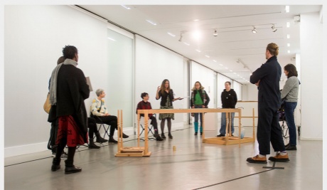 A crowd of people stand around a structure in the centre of a white gallery space. 