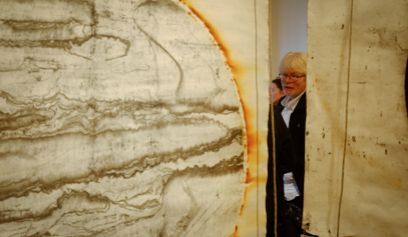 A part image of a large suspended canvas of a partial circle outlined with a smudged orange outline, with sepia wavy lines creating a marble effect inside the circle. Slighty to the right stands Aidan Moesby partially concealed by the canvases.