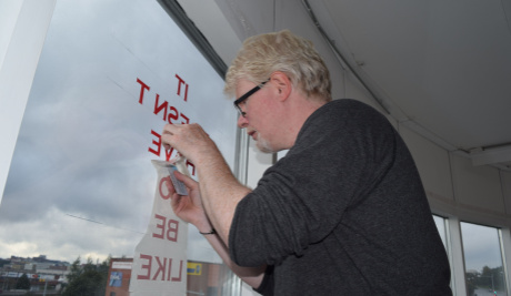 A photograph of Aidan Moesby standing in profile wearing a dark grey jumper and in the process of applying red vinyl lettering to a large window of which only the words 'it', 'be' and 'like' are legible. Through the window, the sky is grey.