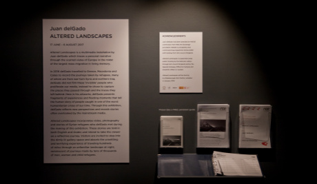 A photograph of the exhibition overview for Altered Landscapes. The overview is printed in black text on a large white board and flyers are attached to the wall next to it.